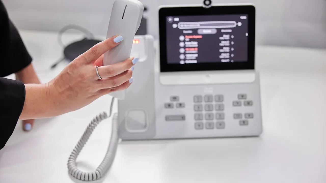 A hand lifts the handset of a white Cisco video phone showing a contacts list on its touchscreen.