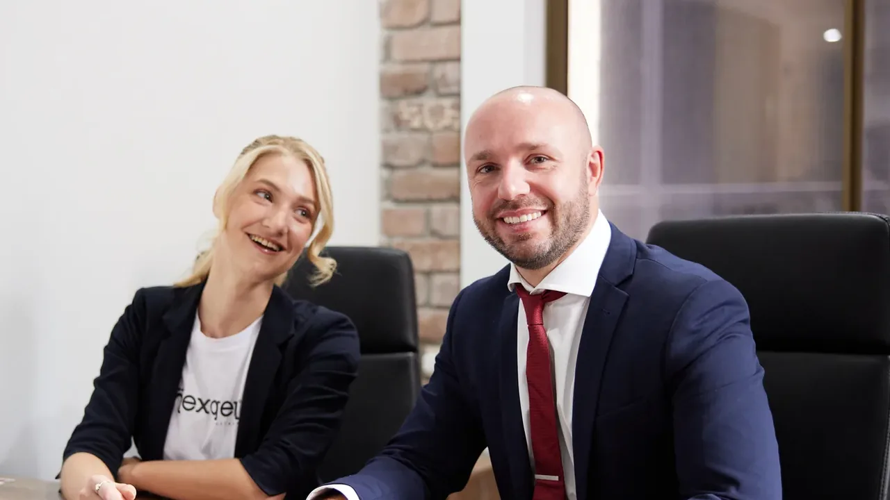 A woman in a NexGen shirt and a man in a navy suit smile together at an office desk with a Cisco desk phone.