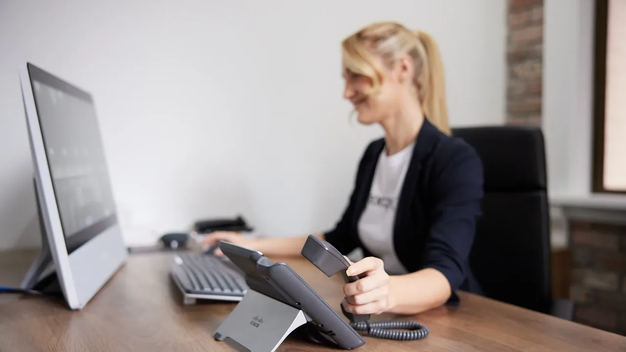 A woman lifts the handset of a Cisco desk phone at her desk, monitor and keyboard beside her.